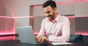 Homem sorrindo usa notebook em ambiente moderno com iluminação rosa neon, sentado à mesa de trabalho em escritório contemporâneo.