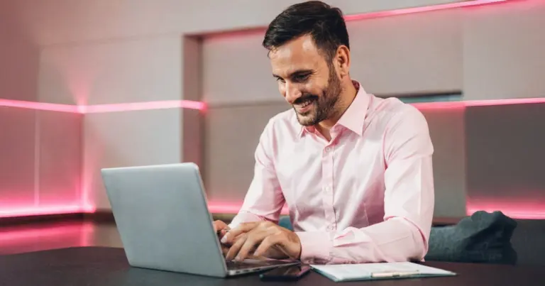 Homem sorrindo usa notebook em ambiente moderno com iluminação rosa neon, sentado à mesa de trabalho em escritório contemporâneo.