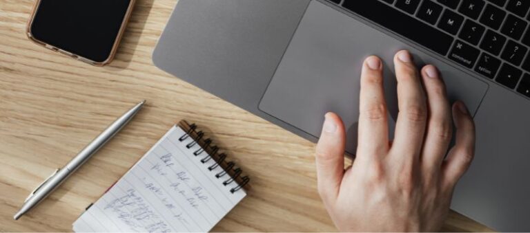 mão em um notebook em uma mesa de madeira ao lado de um bloco de notas, uma caneta e um celular
